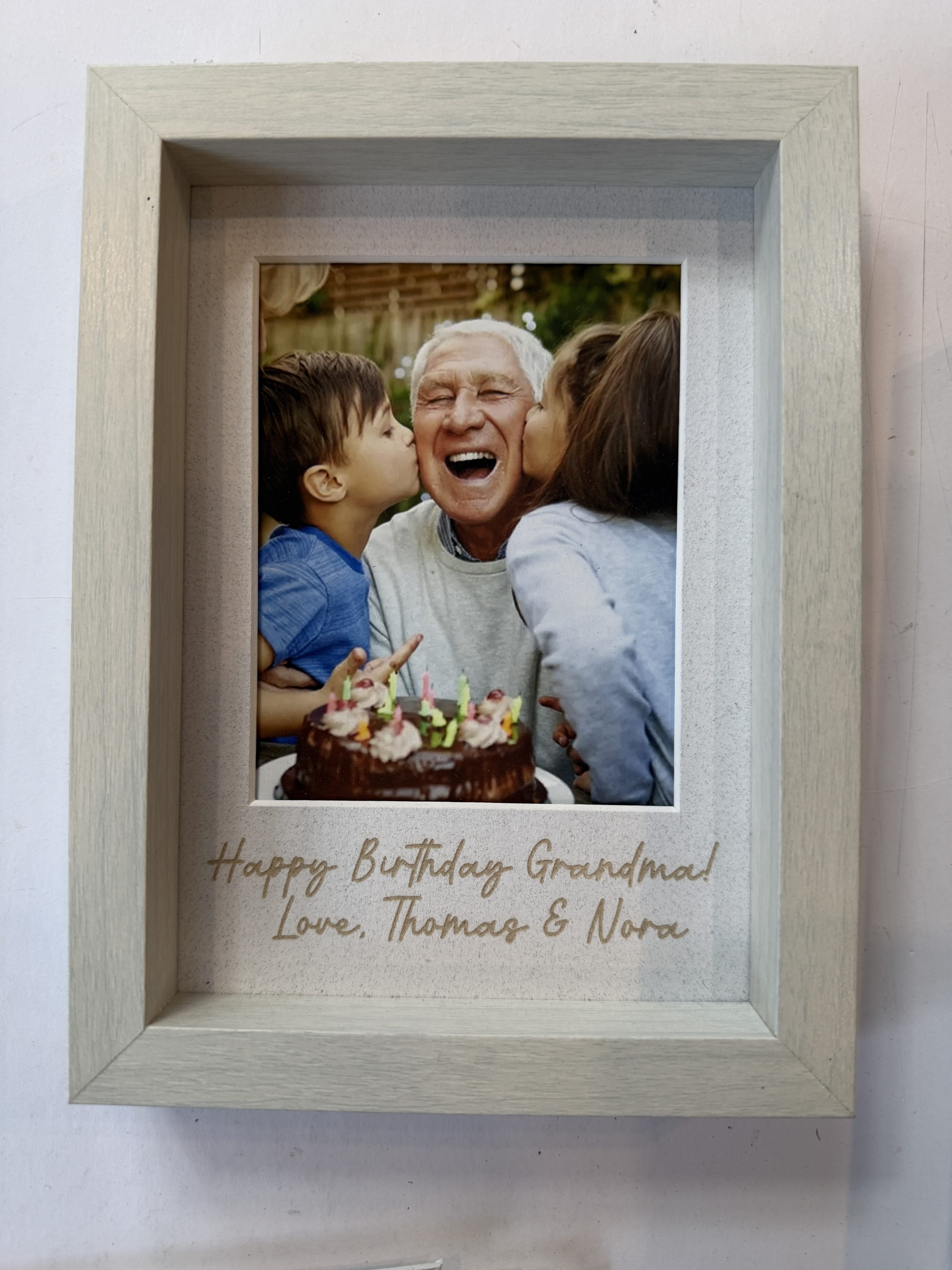 A framed photo of a happy elderly man being kissed on the cheeks by two small children, with a birthday cake visible in front, accompanied by a message that reads 'Happy Birthday Grandma! Love, Thomas & Nora'.